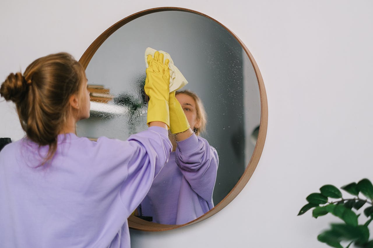 our-story A woman wearing yellow gloves cleans a round wall mirror indoors, reflecting her image.